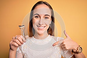 Young beautiful woman using eyelash curler standing over isolated yellow background with surprise face pointing finger to himself