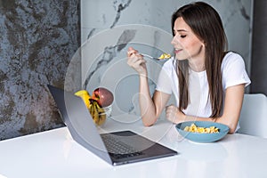 Young beautiful woman using laptop while having breakfast in modern kitchen