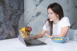 Young beautiful woman using laptop while having breakfast in modern kitchen