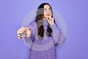 Young beautiful woman using television remote control over isolated purple background serious face thinking about question, very