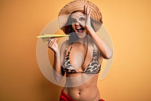 Young beautiful woman on vacation wearing bikini and summer hat holding paper airplane with happy face smiling doing ok sign with