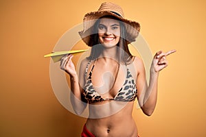 Young beautiful woman on vacation wearing bikini and summer hat holding paper airplane very happy pointing with hand and finger to