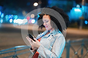 Young beautiful woman with very curly afro hair using mobile phone at night illuminated street. Unusual trendy girl with