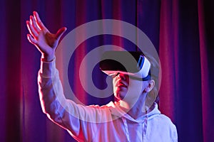 Young beautiful woman in virtual reality glasses and a white hooded sweater on a blue-violet background. Modern technologies