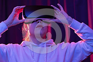 Young beautiful woman in virtual reality glasses and a white hooded sweater on a blue-violet background. Modern technologies
