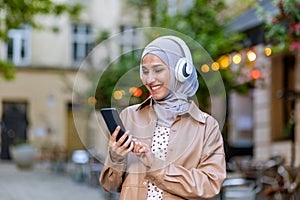 Young beautiful woman walking in the city, Muslim woman in hijab on evening walk in headphones listening to music