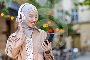 Young beautiful woman walking in the city, Muslim woman in hijab on evening walk in headphones listening to music