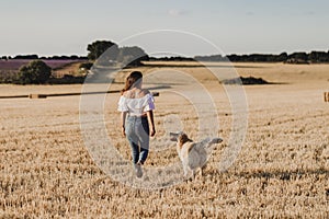 Young beautiful woman walking with her golden retriever dog on a yellow field at sunset. Nature and lifestyle outdoors