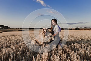 Young beautiful woman walking with her golden retriever dog on a yellow field at sunset. Nature and lifestyle outdoors