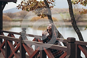 Young beautiful woman in a warm classic coat drinking tea near lake