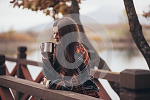 Young beautiful woman in a warm classic coat drinking tea near lake