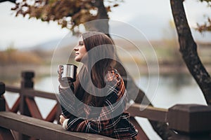 Young beautiful woman in a warm classic coat drinking tea near lake