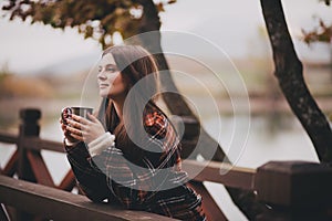 Young beautiful woman in a warm classic coat drinking tea near lake