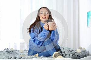 Young beautiful woman in warm sweater with cup of hot drink on bed