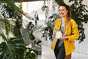 Young beautiful woman watering flowers and plants in her home garden or in a greenhouse, smiling and looking at the camera