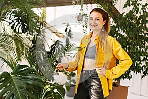 Young beautiful woman watering flowers and plants in her home garden or in a greenhouse, smiling and looking at the camera