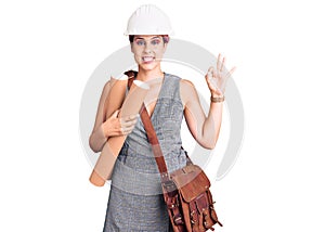 Young beautiful woman wearing architect hardhat and leather bag holding blueprints doing ok sign with fingers, smiling friendly