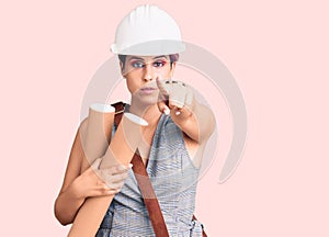 Young beautiful woman wearing architect hardhat and leather bag holding blueprints pointing with finger to the camera and to you,