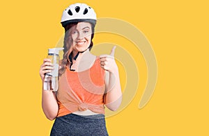 Young beautiful woman wearing bike helmet and holding water bottle smiling happy and positive, thumb up doing excellent and