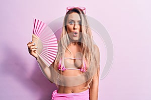 Young beautiful woman wearing bikini and sunglasses using hand fan over pink background scared and amazed with open mouth for