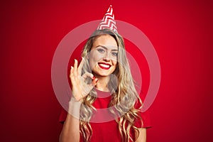 Young beautiful woman wearing bitrhday hat over red isolated background doing ok sign with fingers, excellent symbol