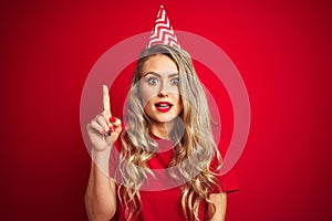 Young beautiful woman wearing bitrhday hat over red isolated background surprised with an idea or question pointing finger with