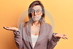 Young beautiful woman wearing business clothes and glasses clueless and confused expression with arms and hands raised