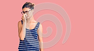 Young beautiful woman wearing casual clothes and glasses smelling something stinky and disgusting, intolerable smell, holding