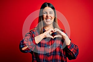 Young beautiful woman wearing casual shirt over red background smiling in love doing heart symbol shape with hands