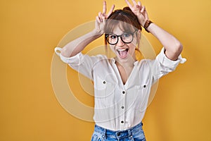 Young beautiful woman wearing casual shirt over yellow background posing funny and crazy with fingers on head as bunny ears,