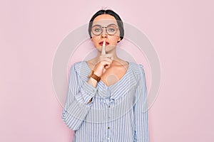 Young beautiful woman wearing casual striped shirt and glasses over pink background asking to be quiet with finger on lips