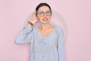 Young beautiful woman wearing casual striped shirt and glasses over pink background smiling with hand over ear listening an