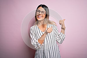 Young beautiful woman wearing casual striped t-shirt and glasses over pink background Pointing to the back behind with hand and