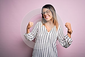 Young beautiful woman wearing casual striped t-shirt and glasses over pink background very happy and excited doing winner gesture