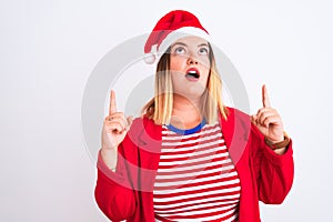 Young beautiful woman wearing Christmas Santa hat over isolated white background amazed and surprised looking up and pointing with