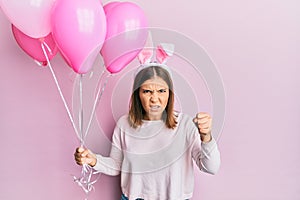 Young beautiful woman wearing cute easter bunny ears and holding balloons annoyed and frustrated shouting with anger, yelling