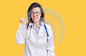 Young beautiful woman wearing doctor stethoscope and glasses angry and mad raising fist frustrated and furious while shouting with