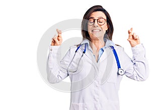 Young beautiful woman wearing doctor stethoscope and glasses gesturing finger crossed smiling with hope and eyes closed