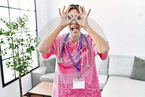 Young beautiful woman wearing doctor uniform and stethoscope doing ok gesture like binoculars sticking tongue out, eyes looking