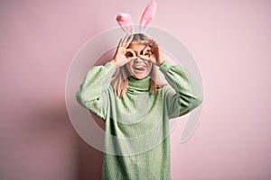 Young beautiful woman wearing easter rabbit ears standing over isolated pink background doing ok gesture like binoculars sticking