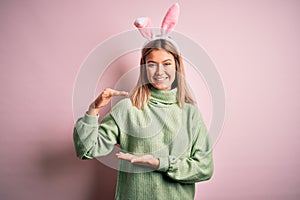 Young beautiful woman wearing easter rabbit ears standing over isolated pink background gesturing with hands showing big and large