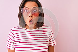 Young beautiful woman wearing fashion sunglasses with hearts over isolated pink background scared in shock with a surprise face,