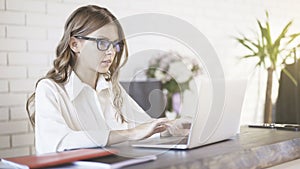 A young beautiful woman wearing glasses typing at her laptop in office. Medium shot