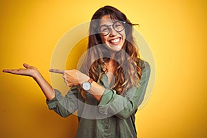 Young beautiful woman wearing green shirt and glasses over yelllow isolated background amazed and smiling to the camera while