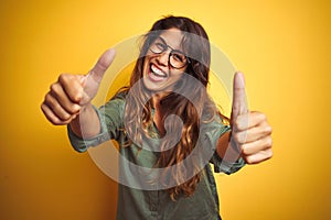 Young beautiful woman wearing green shirt and glasses over yelllow isolated background approving doing positive gesture with hand,