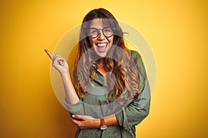 Young beautiful woman wearing green shirt and glasses over yelllow isolated background with a big smile on face, pointing with