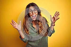 Young beautiful woman wearing green shirt and glasses over yelllow isolated background celebrating crazy and amazed for success