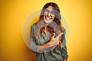 Young beautiful woman wearing green shirt and glasses over yelllow isolated background cheerful with a smile of face pointing with