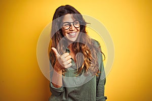 Young beautiful woman wearing green shirt and glasses over yelllow isolated background doing happy thumbs up gesture with hand