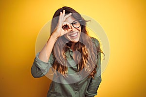 Young beautiful woman wearing green shirt and glasses over yelllow isolated background doing ok gesture with hand smiling, eye